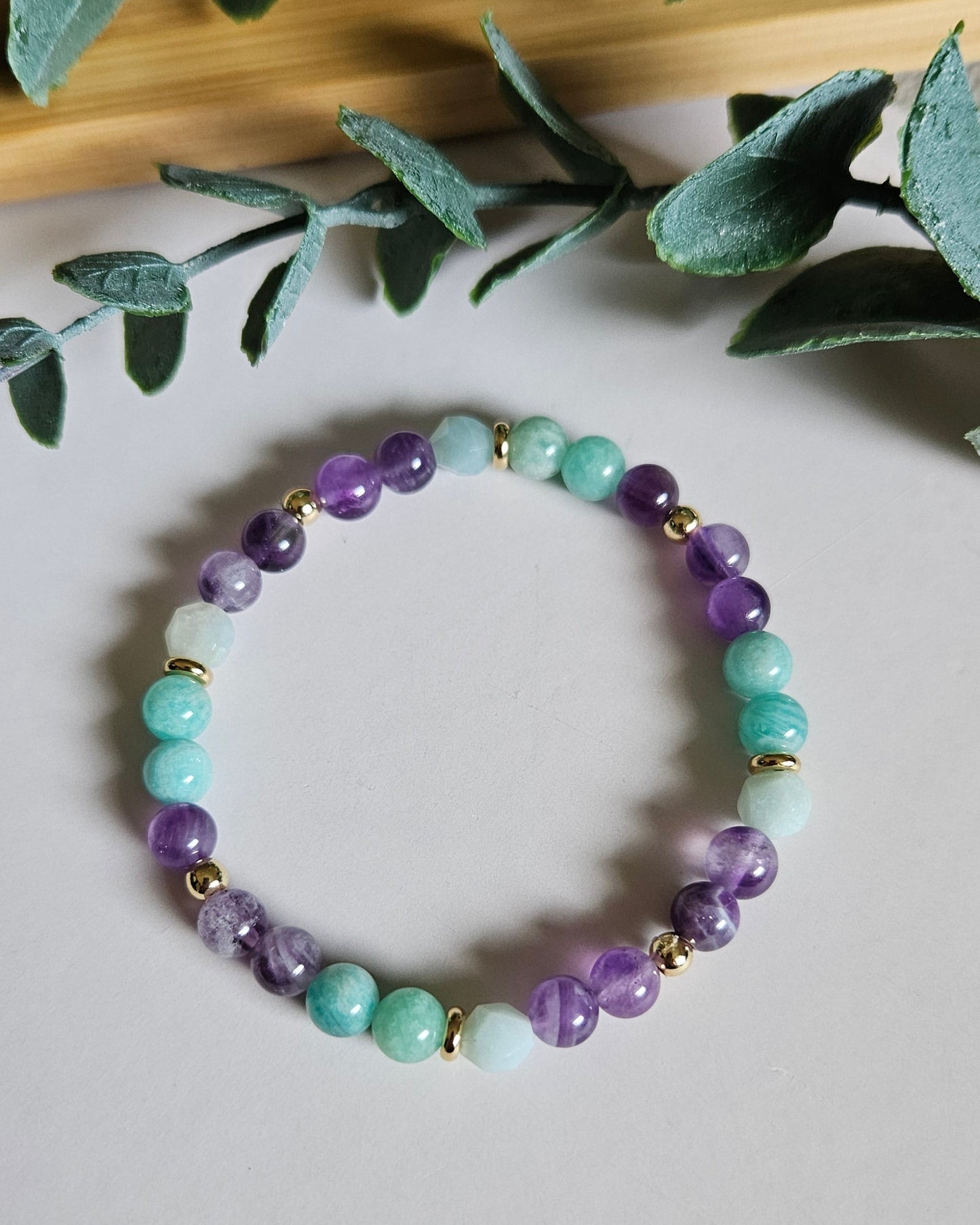 Beaded bracelet with green, purple, and gold beads on a light surface with leaves in the background