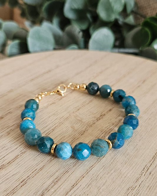 Blue beaded bracelet with gold accents on a wooden surface with greenery in the background