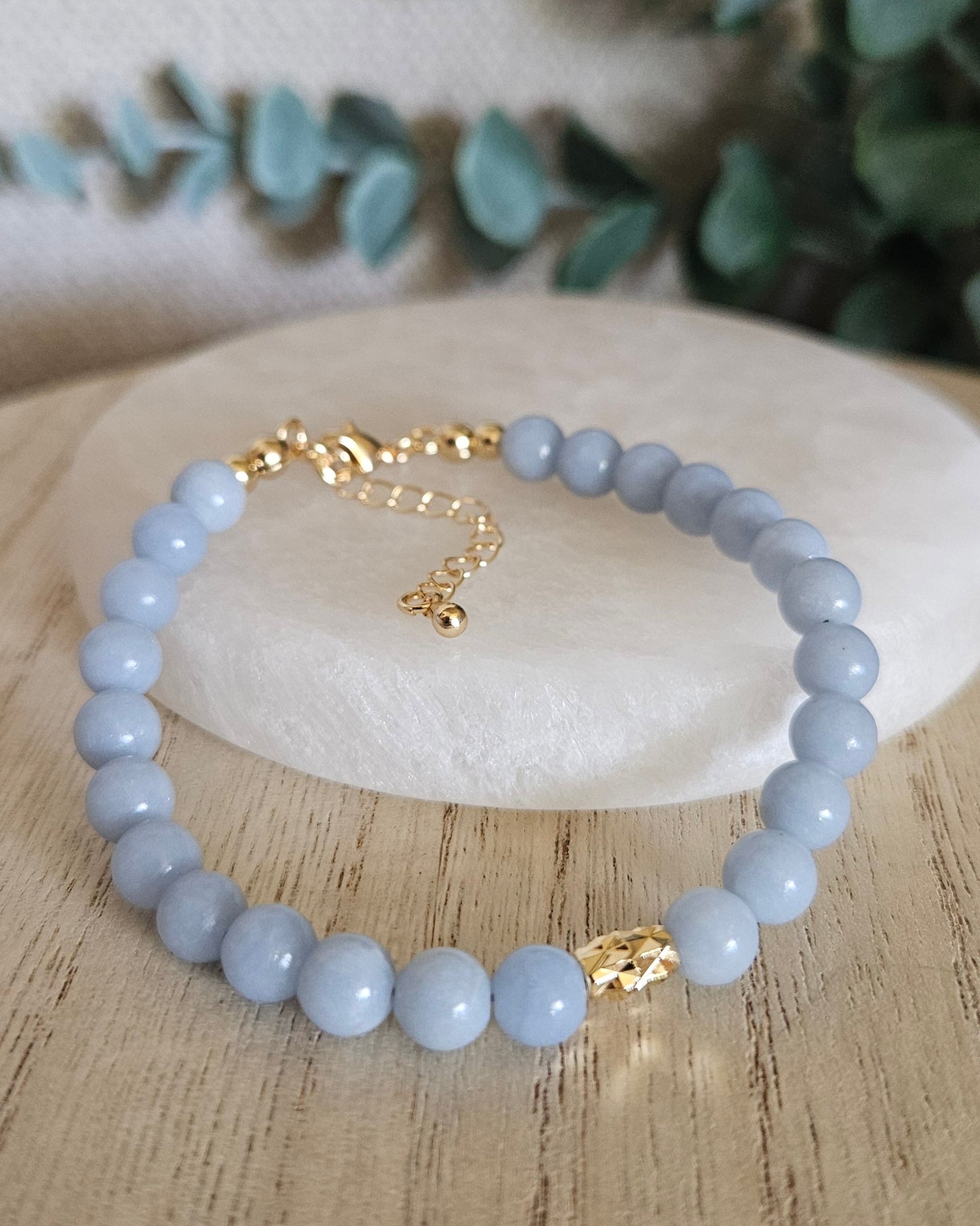 Light blue beaded bracelet on a white stone with gold chain, surrounded by greenery on a wooden surface.