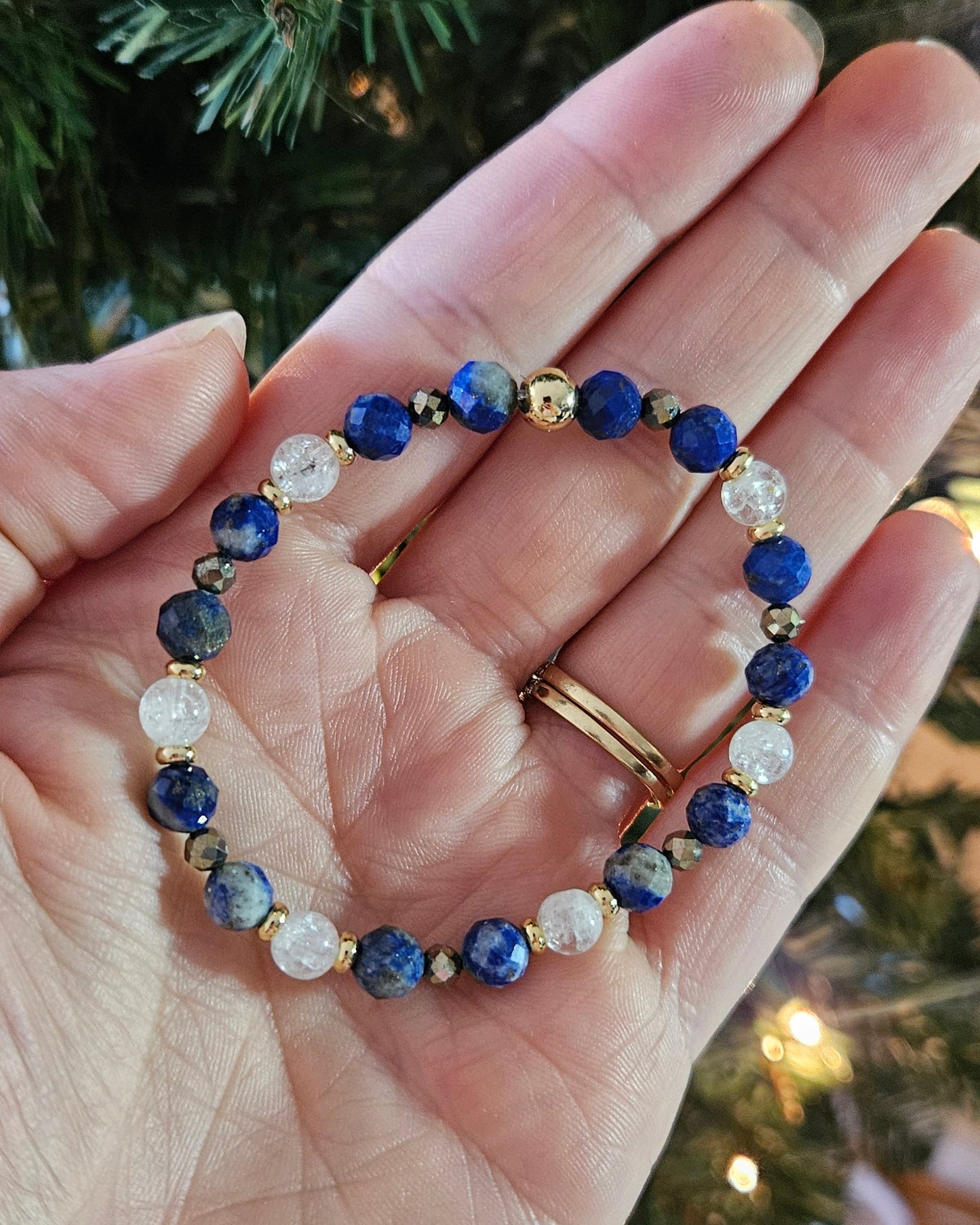 Hand holding a bracelet with blue and gold beads against a blurred Christmas tree background