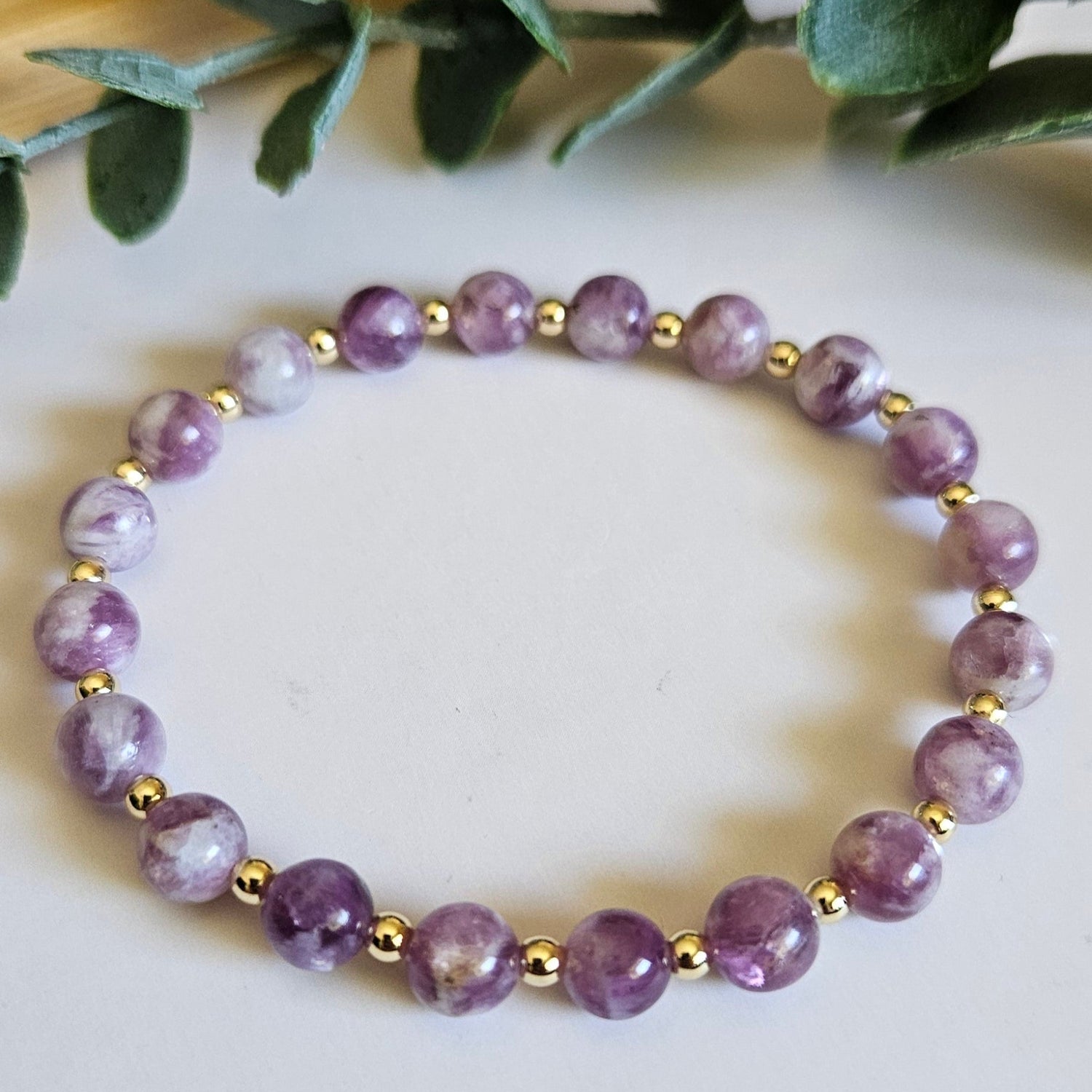 Purple beaded bracelet on a white surface with green leaves in the background