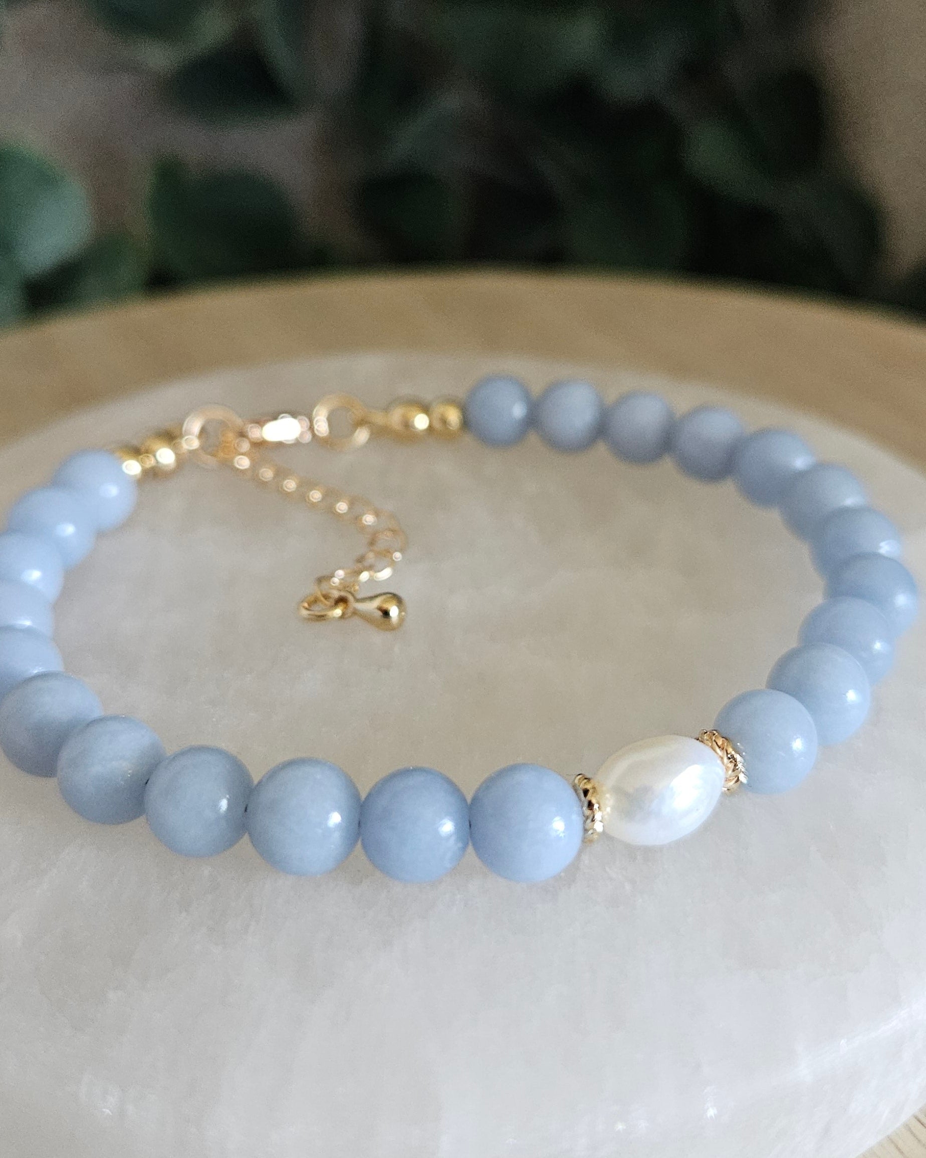 Light blue beaded bracelet on a white dish with a gold chain, against a blurred natural background.