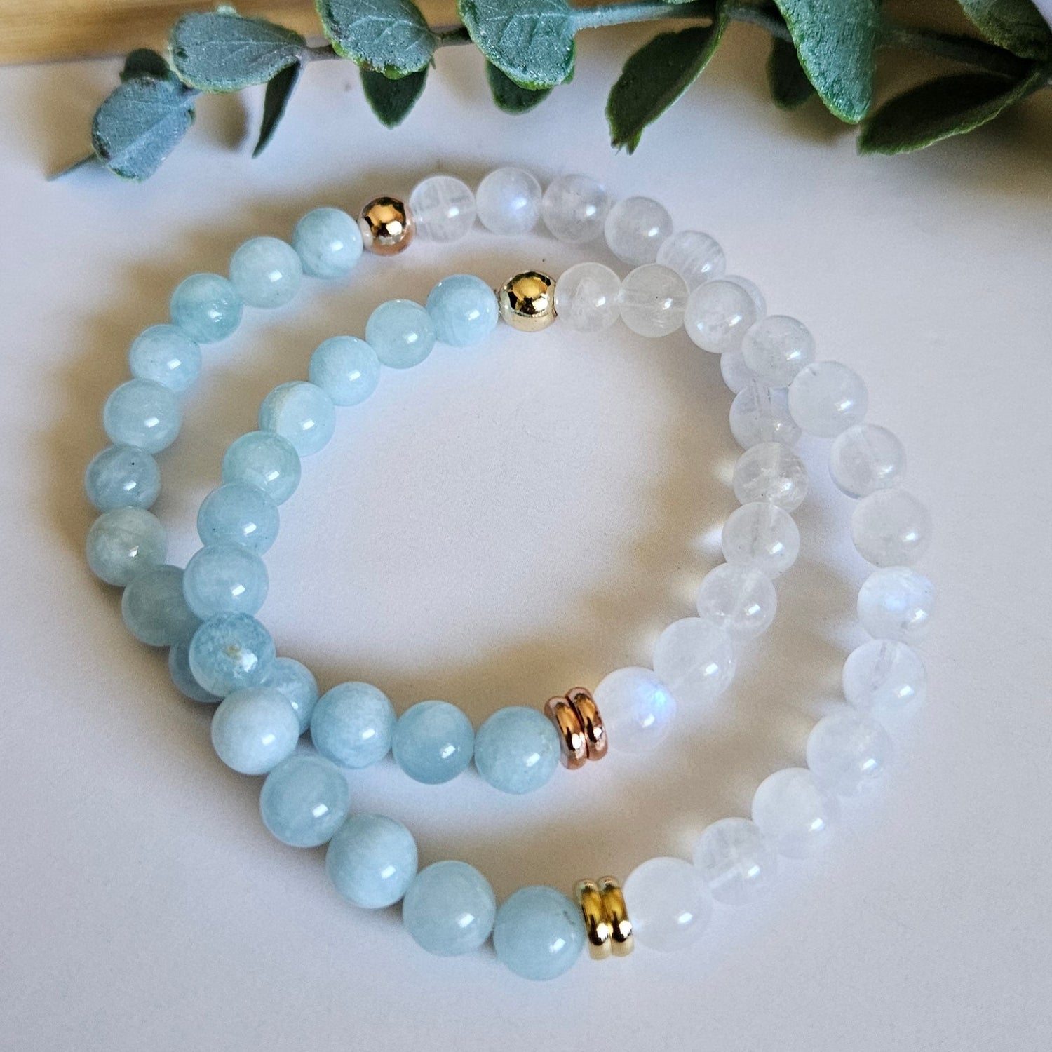 Stack of light blue beaded bracelets with gold accents on a white surface with greenery.