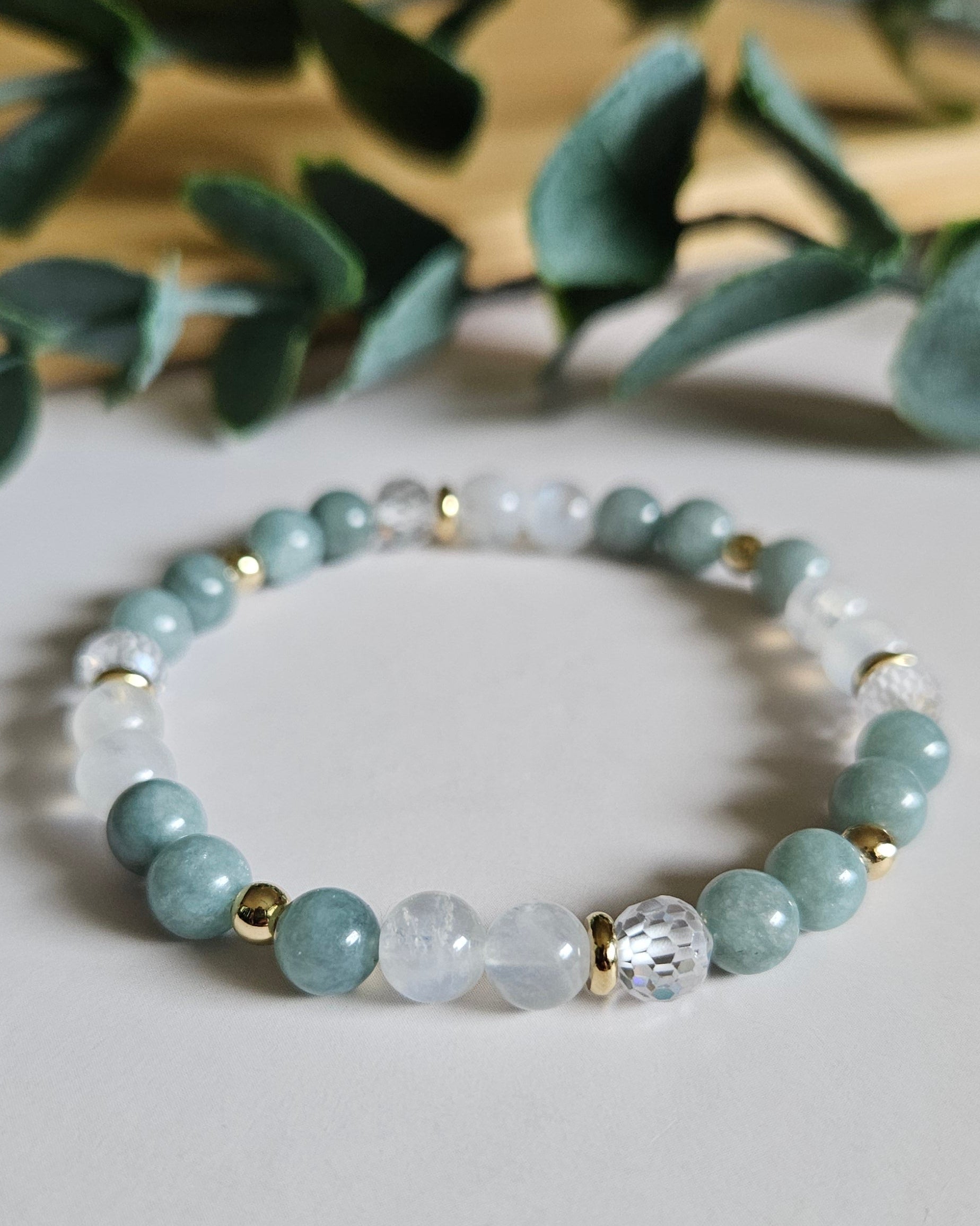Beaded bracelet with light blue and clear beads on a white surface with green leaves in the background