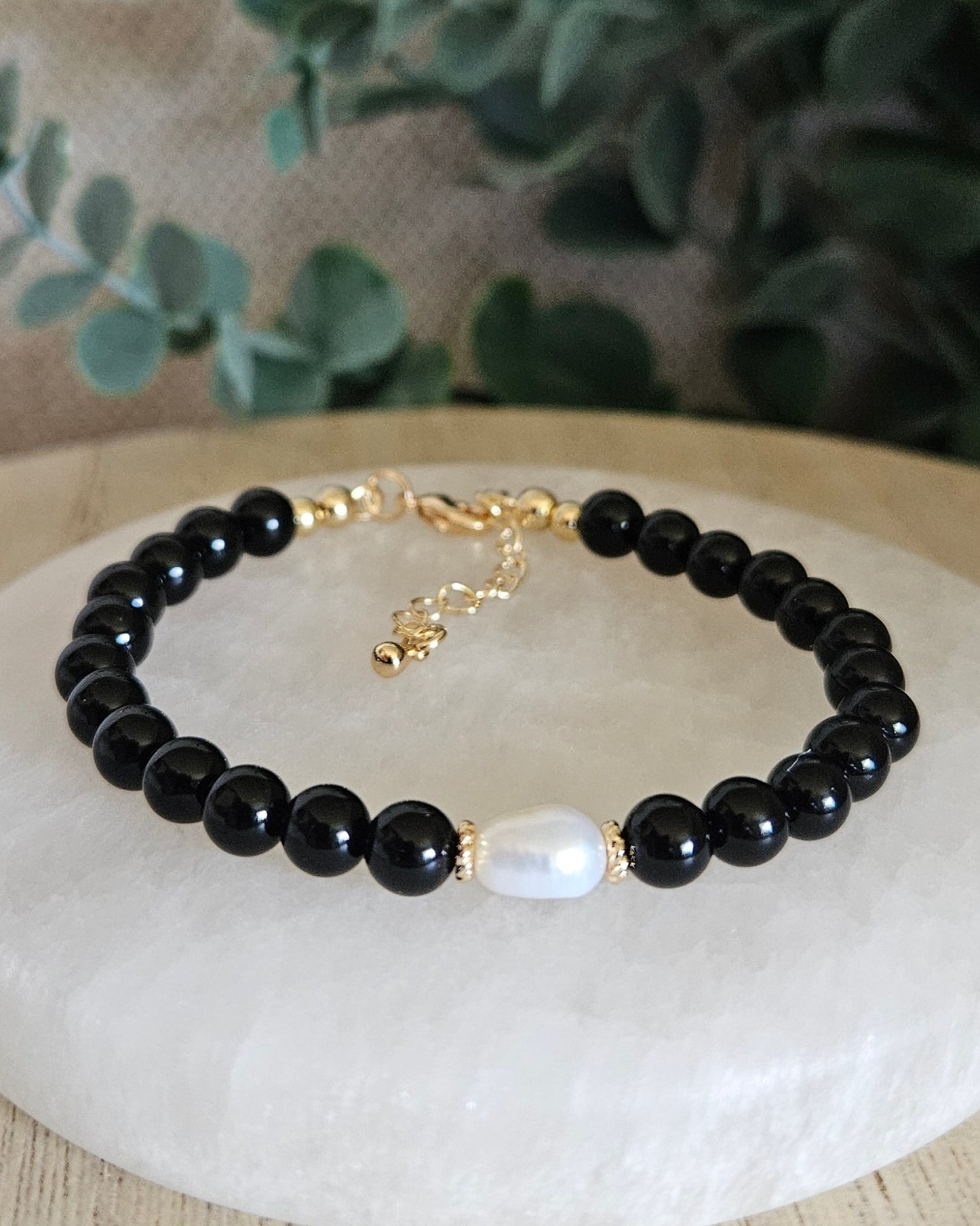 Black beaded bracelet with a gold clasp on a white stone surface, surrounded by greenery.