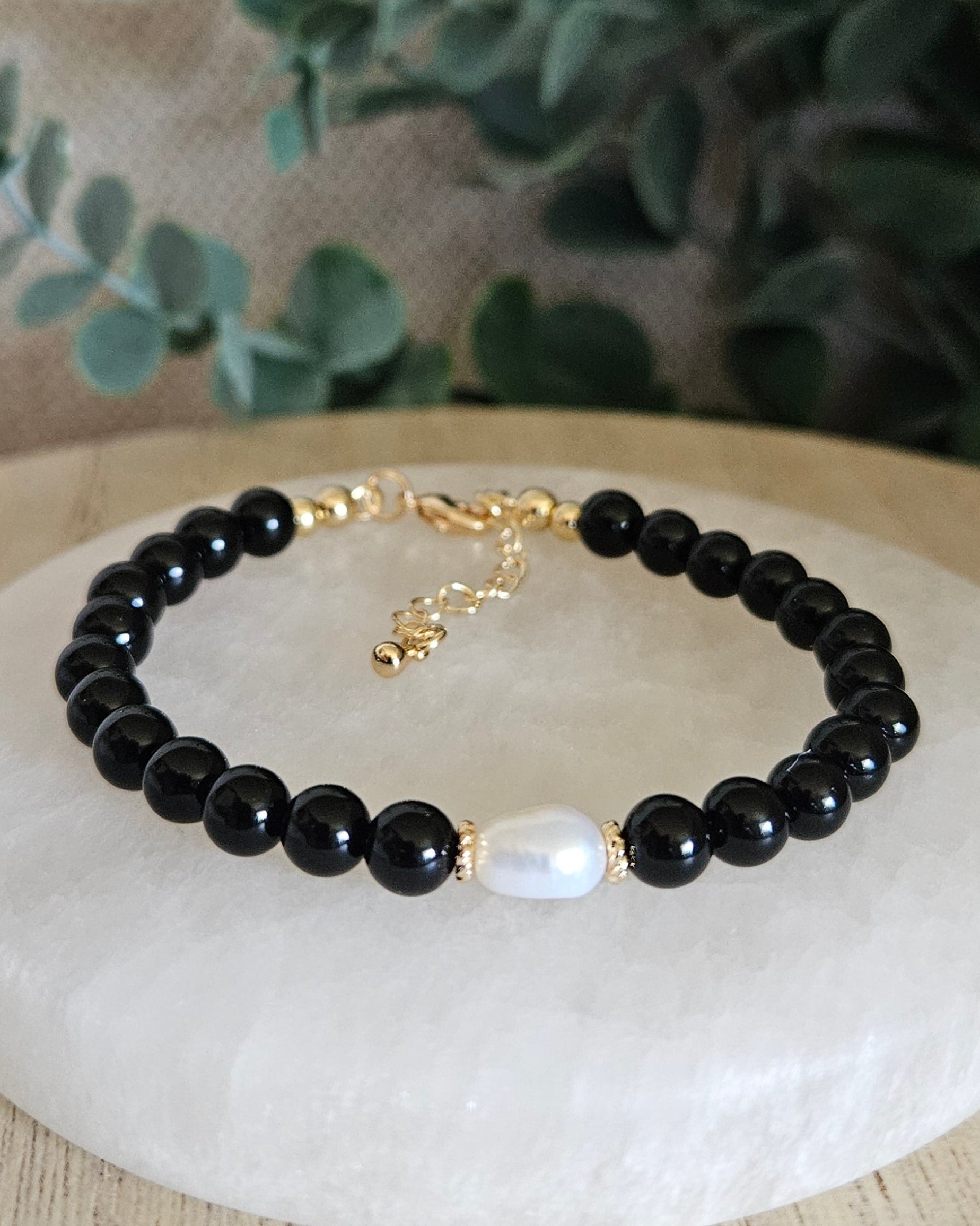 Black beaded bracelet with a gold clasp on a white stone surface, surrounded by greenery.