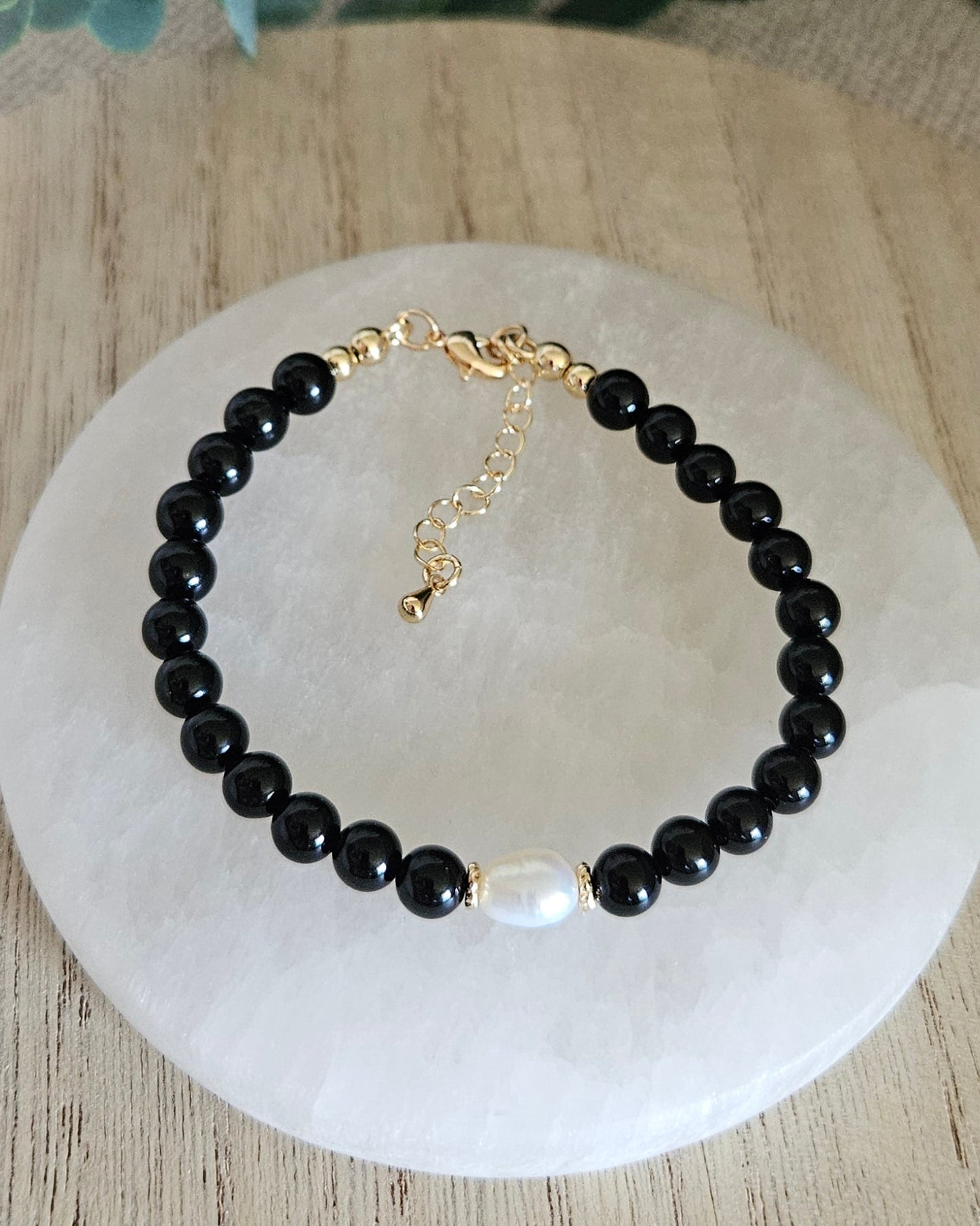Black beaded bracelet with a gold clasp on a white stone surface, surrounded by eucalyptus leaves.