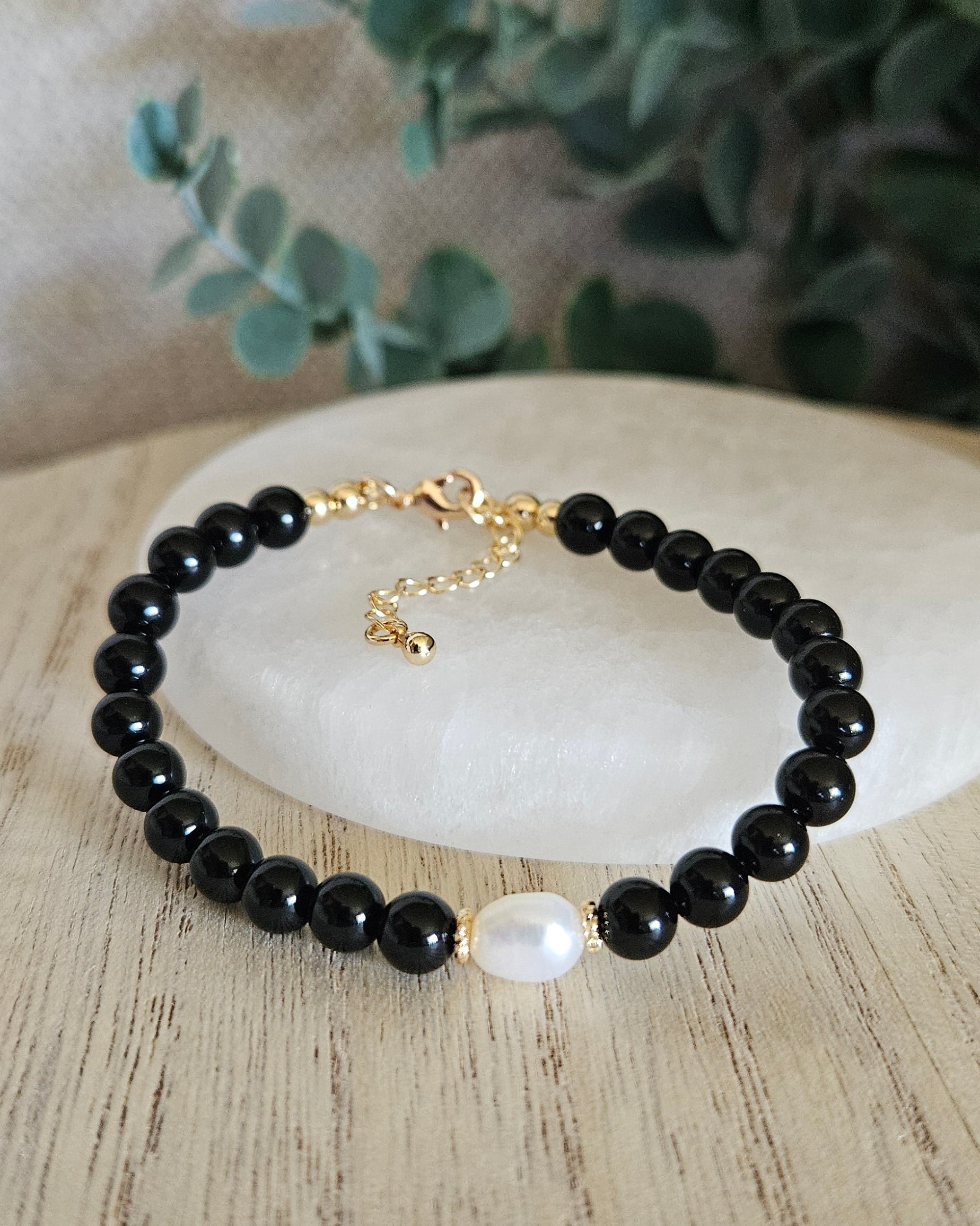 Black beaded bracelet with a gold chain on a white stone with green leaves in the background