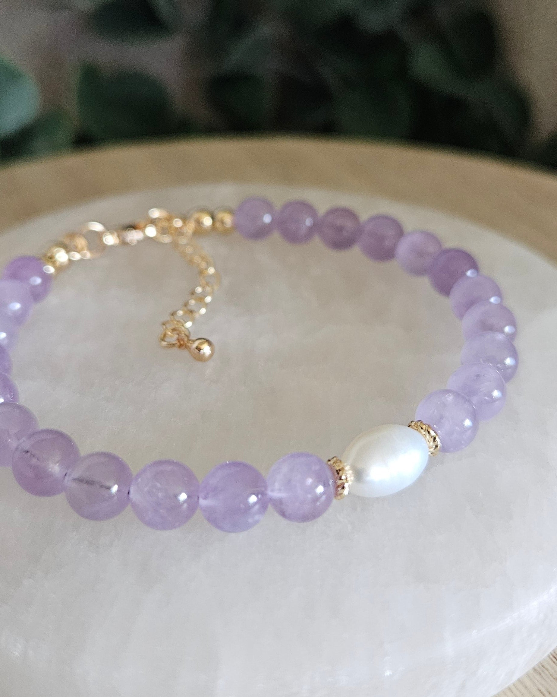 Purple beaded bracelet with a white pearl on a selenite plate, with greenery in the background.